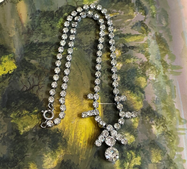 May include: A silver necklace with a large, clear rhinestone pendant. The necklace is made of a chain of small, clear rhinestones.