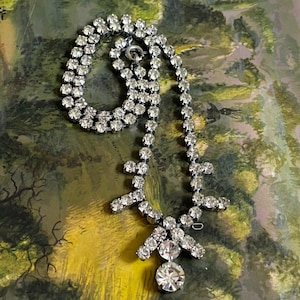 May include: A silver rhinestone necklace with a teardrop pendant. The necklace is made of two rows of rhinestones and has a simple clasp.