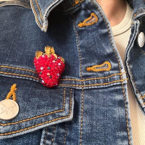May include: A red strawberry brooch with gold accents and a stem, encrusted with sparkling red crystals. The brooch is pinned to a dark blue denim jacket with gold stitching and a silver button.