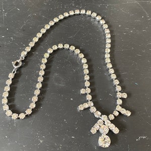 May include: A silver necklace with a large, clear rhinestone pendant. The necklace is made of a chain of small, clear rhinestones.