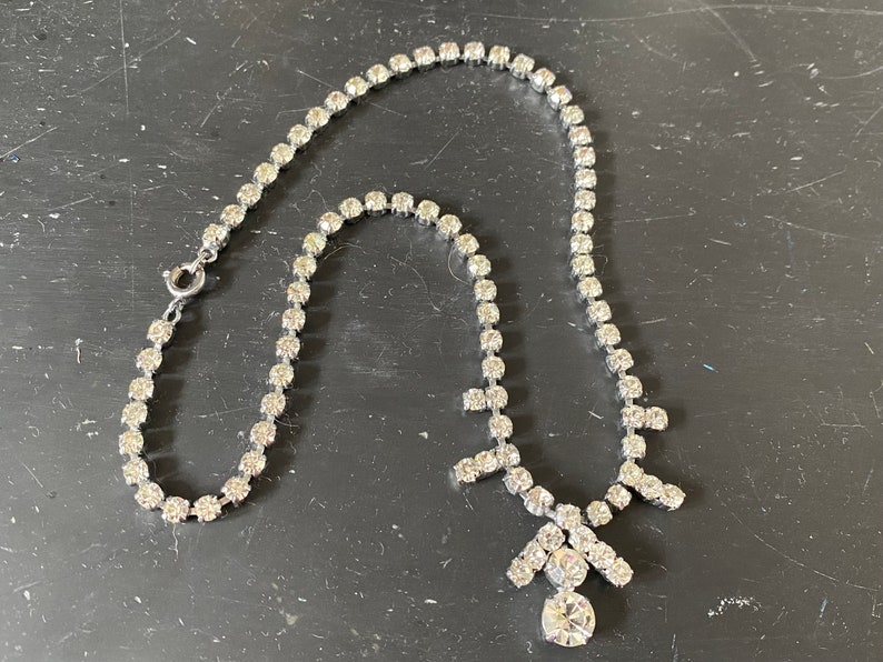 May include: A silver necklace with a large, clear rhinestone pendant. The necklace is made of a chain of small, clear rhinestones.