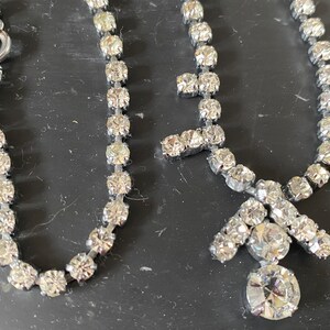 May include: Two silver necklaces with clear rhinestone accents. One necklace is a simple chain with a clasp closure. The other necklace has a pendant with a large rhinestone in the center.