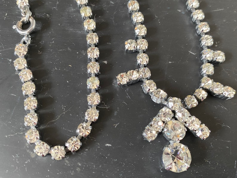 May include: Two silver necklaces with clear rhinestone accents. One necklace is a simple chain with a clasp closure. The other necklace has a pendant with a large rhinestone in the center.
