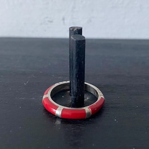 May include: A silver ring featuring a red enamel band with silver rectangular details. The ring is presented on a black stand, set against a neutral backdrop. The design of the ring is simple and elegant.