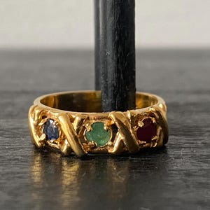 May include: A gold ring featuring three gemstones in blue, green, and red. The ring has an ornate design with an X-shaped pattern between the gemstones. The ring is displayed on a black stand.