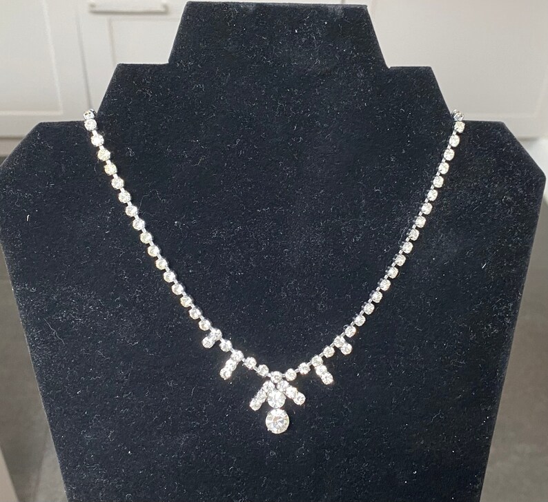 May include: A silver necklace with a teardrop-shaped pendant adorned with clear rhinestones. The necklace is displayed on a black velvet background.