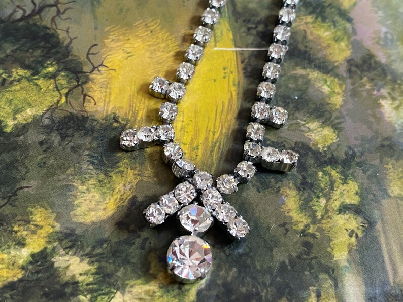 May include: A silver rhinestone necklace with a large, round, faceted rhinestone pendant. The necklace has a delicate, feminine design and is perfect for a special occasion.