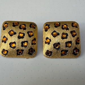 May include: A pair of gold-toned square clip-on earrings. Each earring features a textured surface with a pattern of dark spots, each containing a small, amber-colored gem. The earrings are displayed on a white surface.