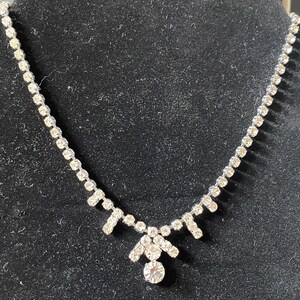 May include: A silver necklace with a teardrop-shaped pendant made of clear rhinestones. The necklace is made of a chain of small rhinestones.