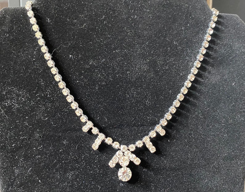 May include: A silver necklace with a teardrop-shaped pendant made of clear rhinestones. The necklace is made of a chain of small rhinestones.