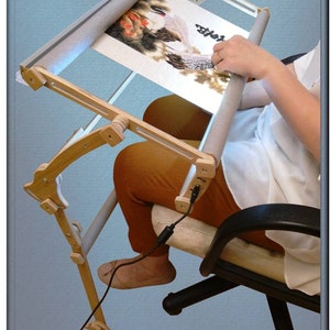 Puede incluir: Un soporte de bordado de madera con una tela de color claro estirada sobre &eacute;l. El soporte est&aacute; unido a una silla y tiene un cable enchufado. El texto "Upheaval on 180&deg;" es visible en la parte inferior de la imagen.