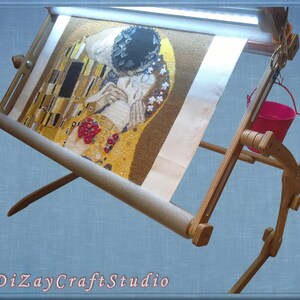 Puede incluir: Un soporte de bordado de madera con una tela de color claro estirada sobre &eacute;l. La tela tiene una imagen colorida de una pintura. El soporte tiene una luz incorporada y un peque&ntilde;o cubo rosa colgando del lado.