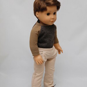 May include: A doll wearing a brown and grey long-sleeved shirt, beige trousers, and brown shoes.