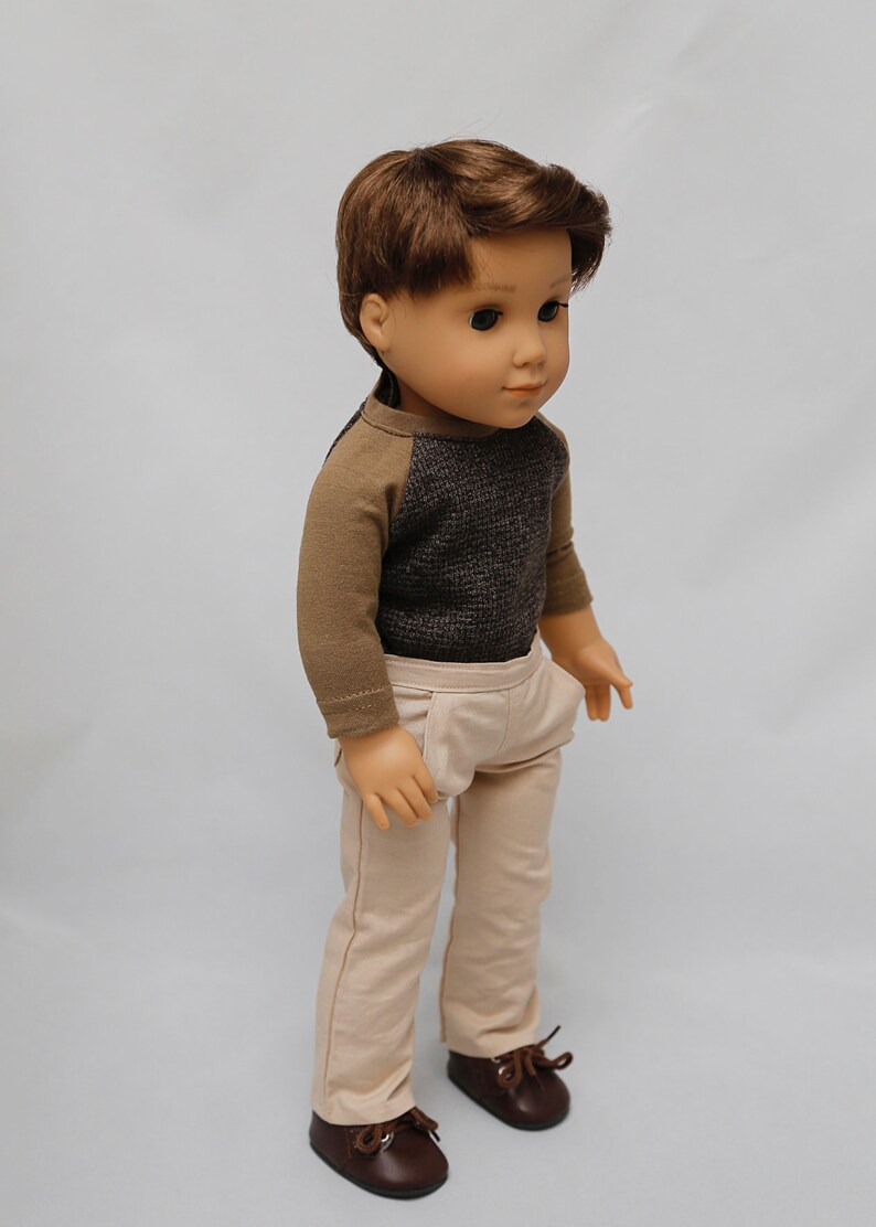May include: A doll wearing a brown and grey long-sleeved shirt, beige trousers, and brown shoes.