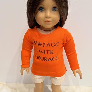 May include: Orange long-sleeved shirt with the text "Voyage with Courage" in brown lettering. The shirt is being worn by a doll with brown hair and blue eyes.
