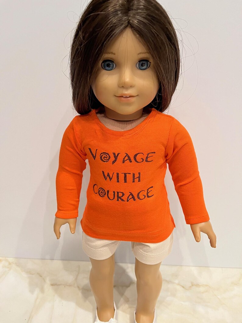 May include: Orange long-sleeved shirt with the text "Voyage with Courage" in brown lettering. The shirt is being worn by a doll with brown hair and blue eyes.