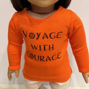 May include: Orange long-sleeved shirt with the text "Voyage with Courage" in black glitter lettering. The shirt is worn by a doll.