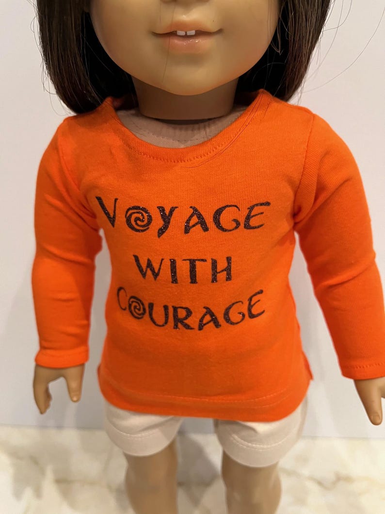 May include: Orange long-sleeved shirt with the text "Voyage with Courage" in black glitter lettering. The shirt is worn by a doll.