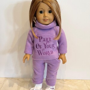 K&ouml;nnte beinhalten: Ein lilafarbenes Fleece-Outfit f&uuml;r eine Puppe, bestehend aus einem Rollkragenpullover mit dem Text "Part of Your World" und einer passenden Hose. Die Puppe tr&auml;gt wei&szlig;e Turnschuhe.