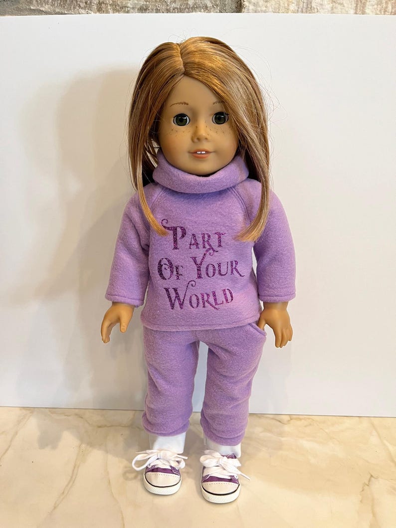 K&ouml;nnte beinhalten: Ein lilafarbenes Fleece-Outfit f&uuml;r eine Puppe, bestehend aus einem Rollkragenpullover mit dem Text "Part of Your World" und einer passenden Hose. Die Puppe tr&auml;gt wei&szlig;e Turnschuhe.