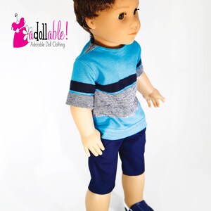 K&ouml;nnte beinhalten: Eine Puppe tr&auml;gt ein blau-graues, gestreiftes T-Shirt mit kurzen &Auml;rmeln, eine dunkelblaue Shorts und blaue Turnschuhe. Die Puppe hat braunes Haar und blaue Augen. Die Puppe steht auf einem wei&szlig;en Hintergrund. "Adorable Doll Clothing" ist in Pink auf wei&szlig;em Hintergrund mit einer rosa Blume geschrieben.