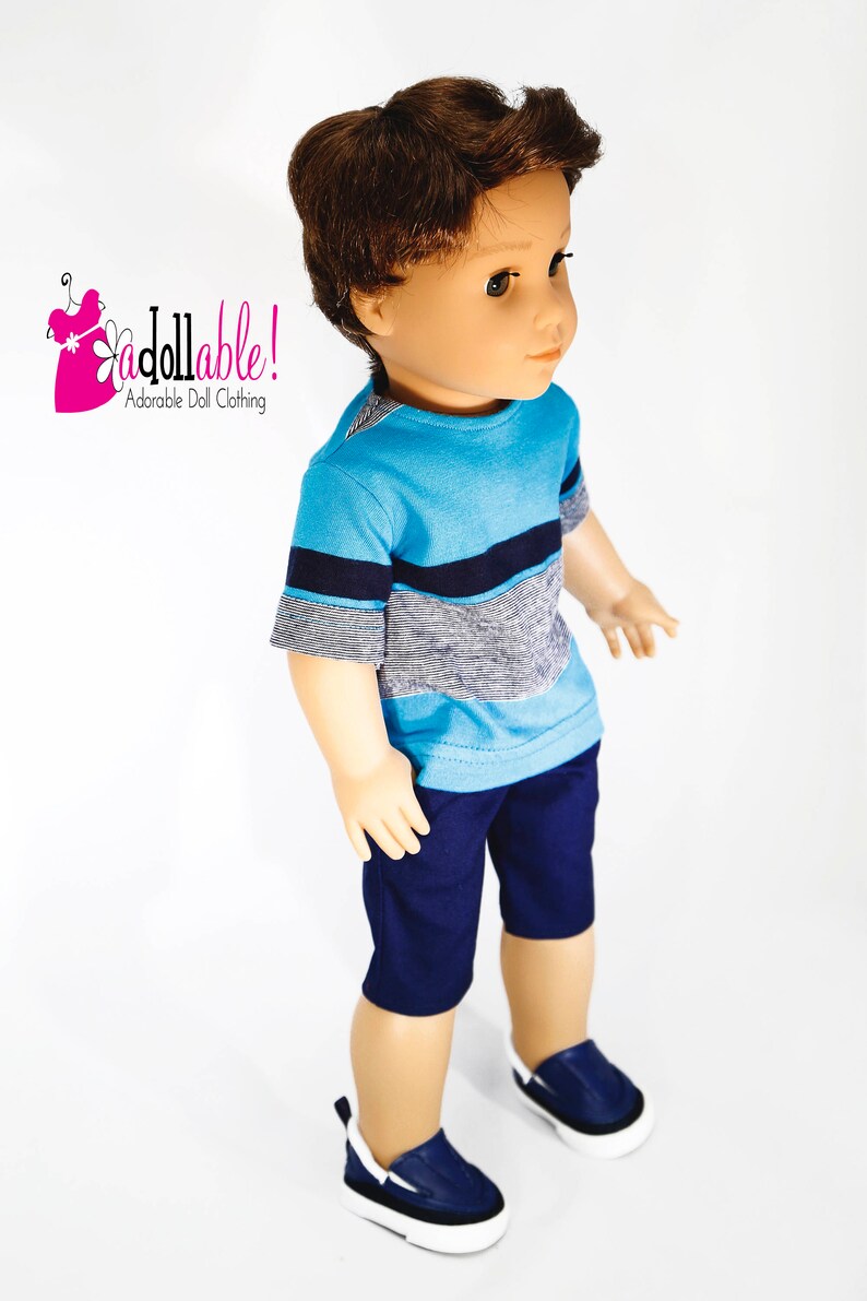 K&ouml;nnte beinhalten: Eine Puppe tr&auml;gt ein blau-graues, gestreiftes T-Shirt mit kurzen &Auml;rmeln, eine dunkelblaue Shorts und blaue Turnschuhe. Die Puppe hat braunes Haar und blaue Augen. Die Puppe steht auf einem wei&szlig;en Hintergrund. "Adorable Doll Clothing" ist in Pink auf wei&szlig;em Hintergrund mit einer rosa Blume geschrieben.