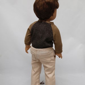 May include: A doll wearing a brown and grey long-sleeved shirt and beige trousers. The doll has brown hair and is standing on a white background.
