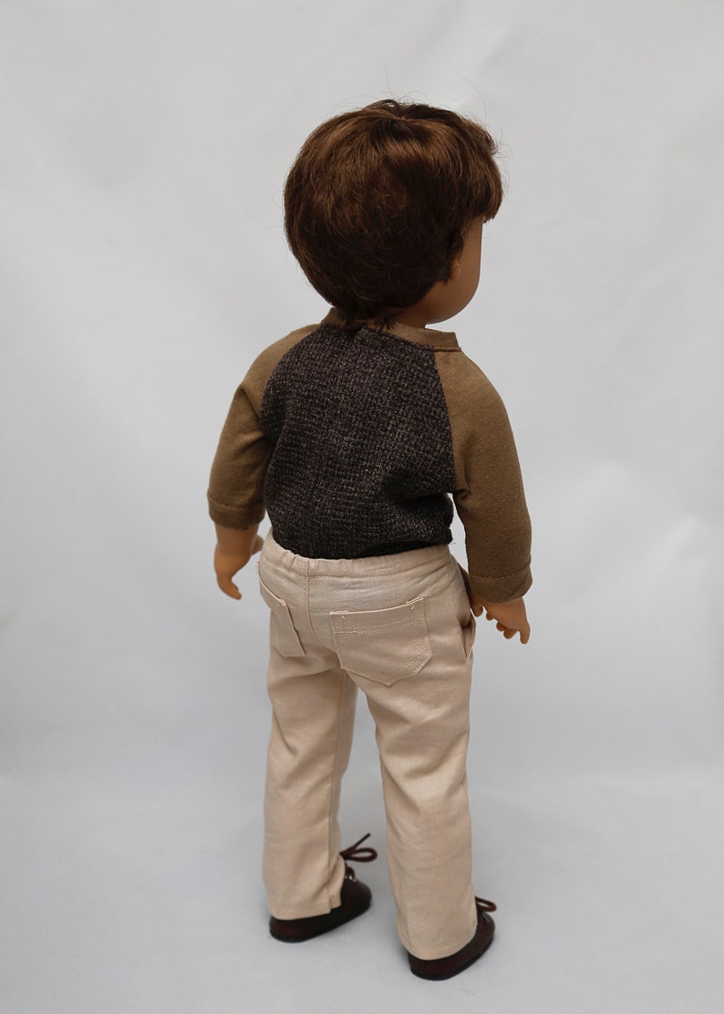 May include: A doll wearing a brown and grey long-sleeved shirt and beige trousers. The doll has brown hair and is standing on a white background.