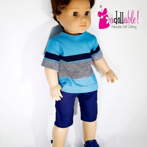 K&ouml;nnte beinhalten: Eine Puppe tr&auml;gt ein blau-wei&szlig; gestreiftes T-Shirt, eine dunkelblaue Shorts und blaue Turnschuhe. Die Puppe hat braunes Haar und blaue Augen. Die Puppe steht auf einem wei&szlig;en Hintergrund. "Jdollable! Adorable Doll Clothing" steht in rosa und schwarzer Schrift in der oberen rechten Ecke des Bildes.