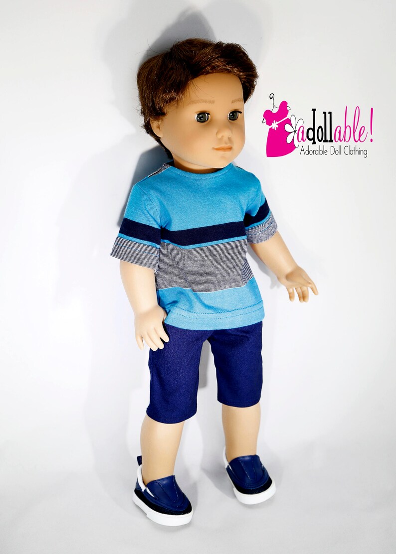 K&ouml;nnte beinhalten: Eine Puppe tr&auml;gt ein blau-wei&szlig; gestreiftes T-Shirt, eine dunkelblaue Shorts und blaue Turnschuhe. Die Puppe hat braunes Haar und blaue Augen. Die Puppe steht auf einem wei&szlig;en Hintergrund. "Jdollable! Adorable Doll Clothing" steht in rosa und schwarzer Schrift in der oberen rechten Ecke des Bildes.