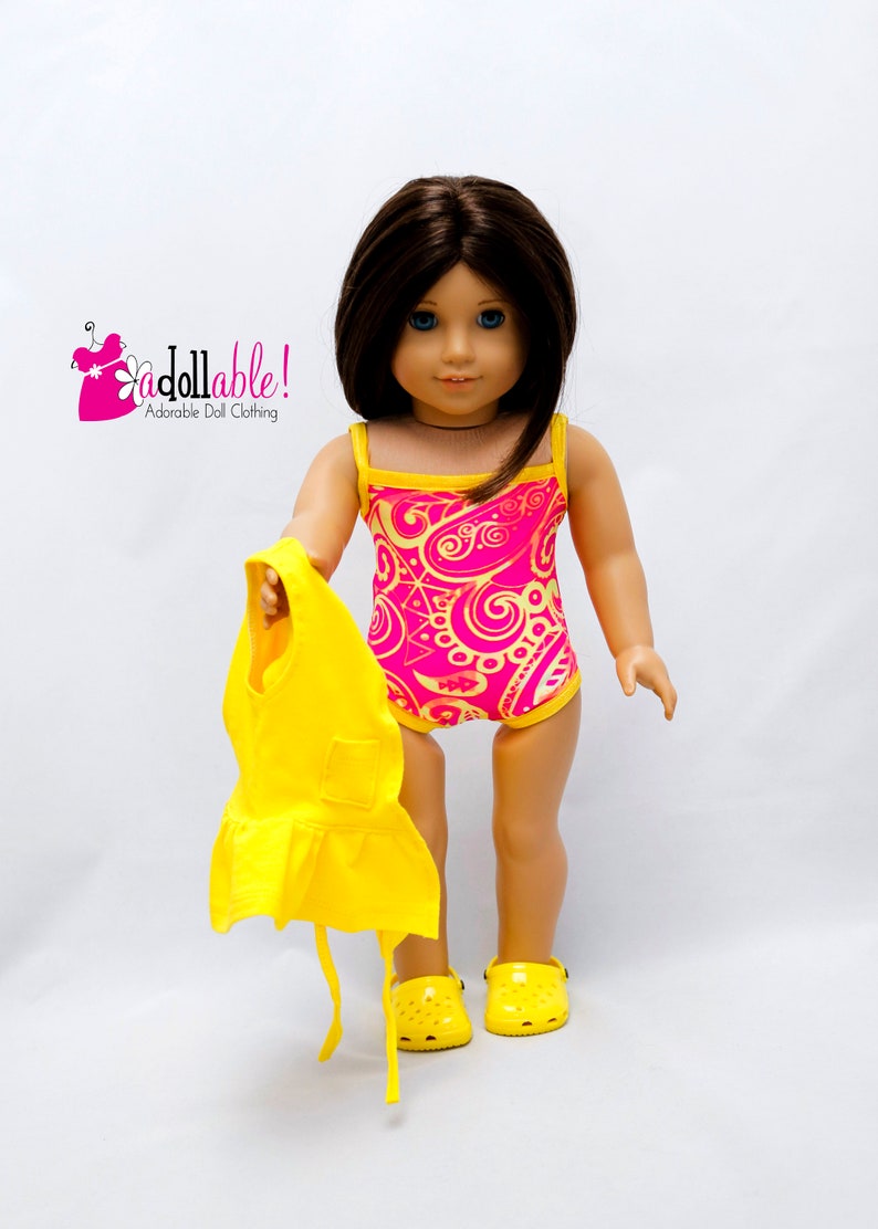 May include: A doll wearing a pink and yellow swimsuit with a paisley pattern. The doll is holding a yellow dress and is wearing yellow shoes.
