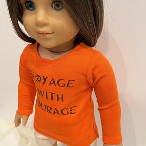 May include: Orange long-sleeved shirt with the text "Voyage with Courage" in black glitter lettering. The shirt is worn by a doll with brown hair and blue eyes.