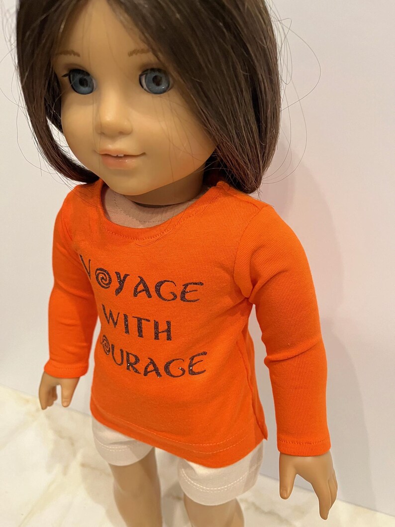 May include: Orange long-sleeved shirt with the text "Voyage with Courage" in black glitter lettering. The shirt is worn by a doll with brown hair and blue eyes.