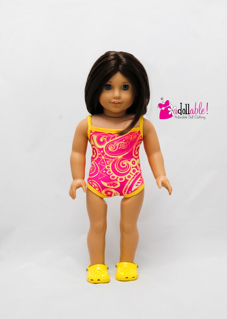 May include: A doll wearing a pink and yellow swimsuit with a paisley pattern. The doll is wearing yellow clogs.