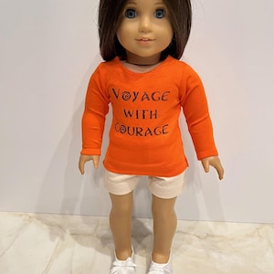 May include: A doll wearing an orange long-sleeved shirt with the text "Voyage with Courage" in brown lettering. The doll is also wearing white shorts and white shoes.