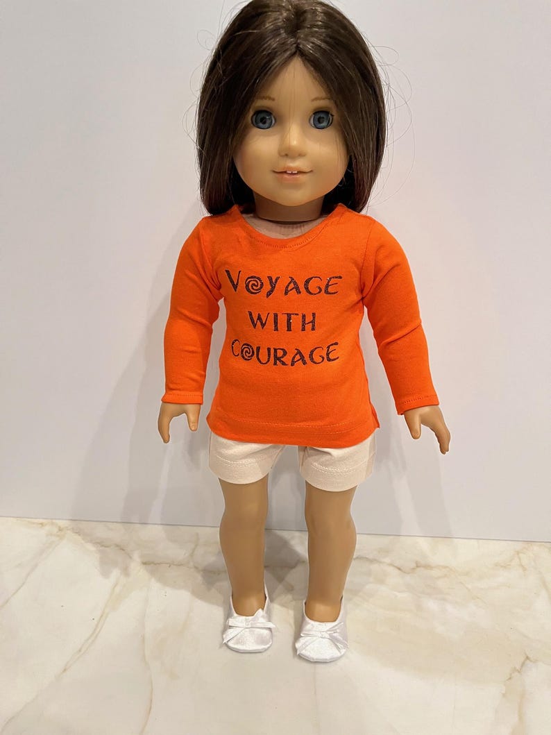 May include: A doll wearing an orange long-sleeved shirt with the text "Voyage with Courage" in brown lettering. The doll is also wearing white shorts and white shoes.