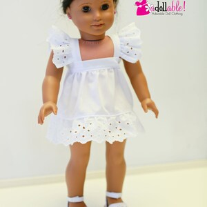 May include: A white eyelet dress with a ruffled hem and cap sleeves. The dress is worn by a doll with brown hair and brown eyes. The doll is wearing white shoes.