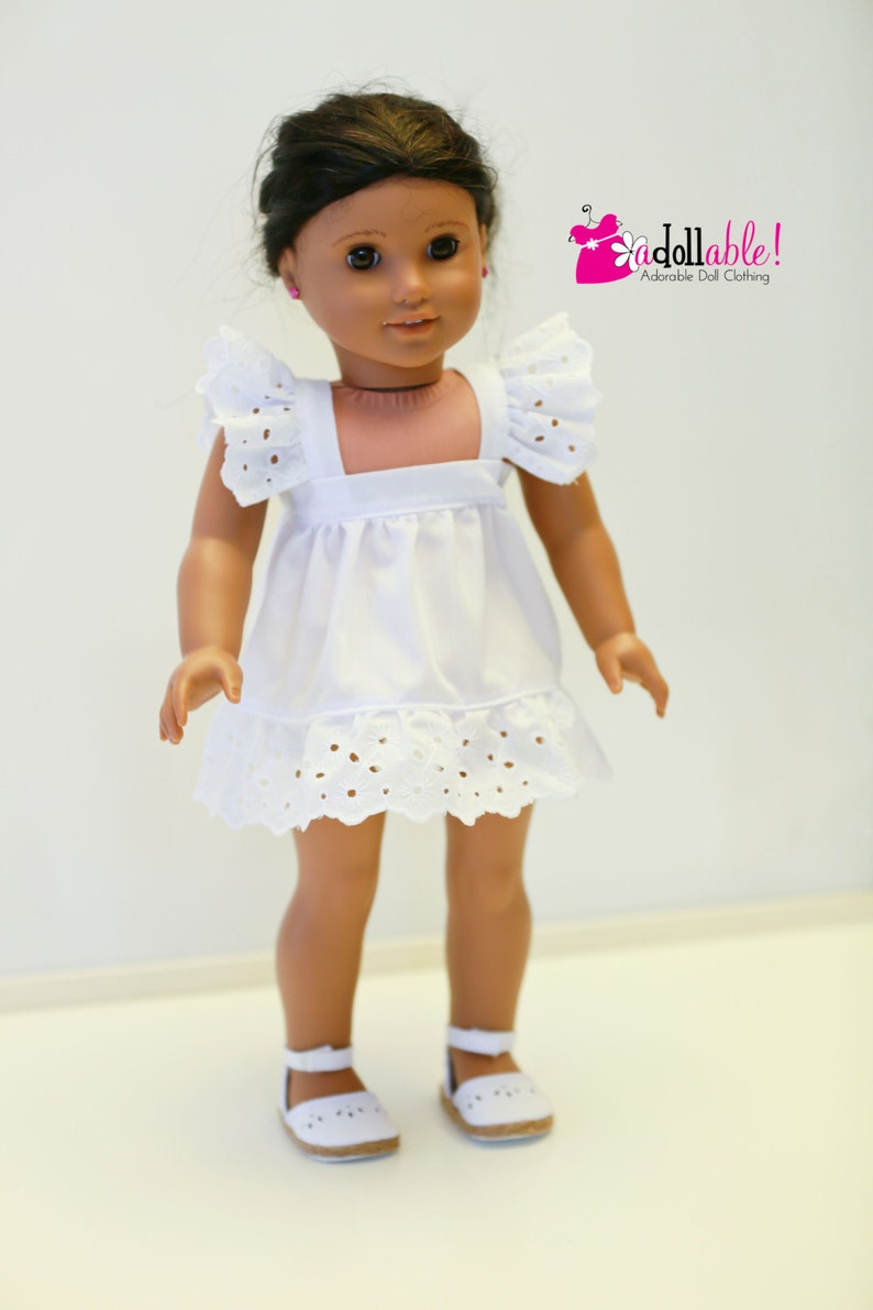 May include: A white eyelet dress with a ruffled hem and cap sleeves. The dress is worn by a doll with brown hair and brown eyes. The doll is wearing white shoes.