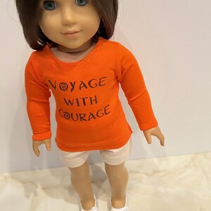 May include: Orange long-sleeved shirt with the text "Voyage with Courage" in brown lettering. The shirt is worn by a doll with brown hair and white shorts. The doll is wearing white shoes.