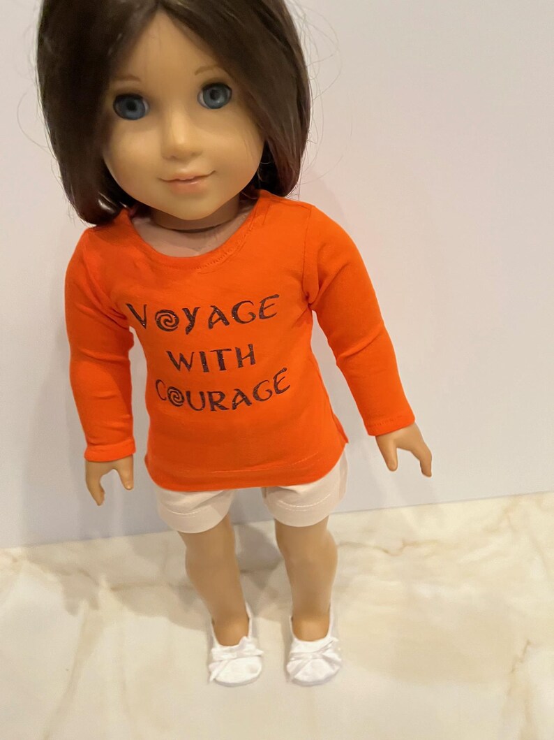 May include: Orange long-sleeved shirt with the text "Voyage with Courage" in brown lettering. The shirt is worn by a doll with brown hair and white shorts. The doll is wearing white shoes.