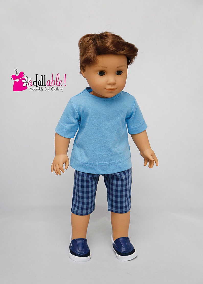 May include: A doll wearing a light blue t-shirt and blue and white checkered shorts. The doll is wearing blue sneakers. The doll has brown hair and blue eyes. The doll is wearing an outfit from "Addollable! Adorable Doll Clothing."