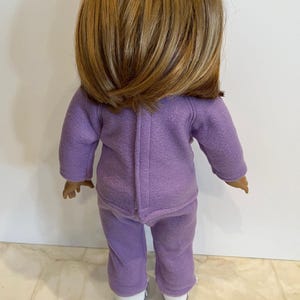 K&ouml;nnte beinhalten: Ein lilafarbener Fleece-Pyjama f&uuml;r eine Puppe. Das Set besteht aus einem lang&auml;rmeligen Oberteil und einer Hose. Die Puppe tr&auml;gt wei&szlig;e Turnschuhe.