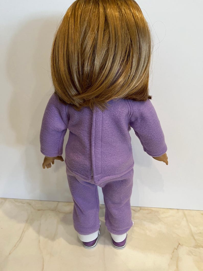 K&ouml;nnte beinhalten: Ein lilafarbener Fleece-Pyjama f&uuml;r eine Puppe. Das Set besteht aus einem lang&auml;rmeligen Oberteil und einer Hose. Die Puppe tr&auml;gt wei&szlig;e Turnschuhe.