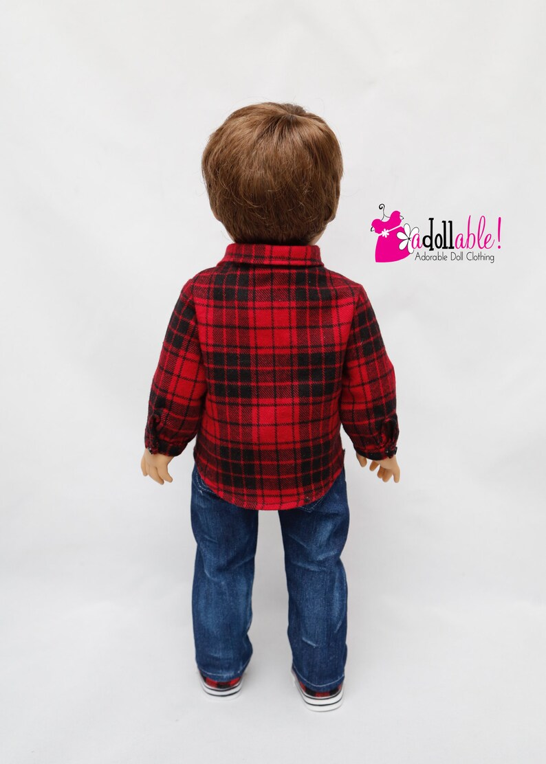 May include: A doll wearing a red and black plaid shirt and blue jeans. The doll has brown hair and is standing on a white background.