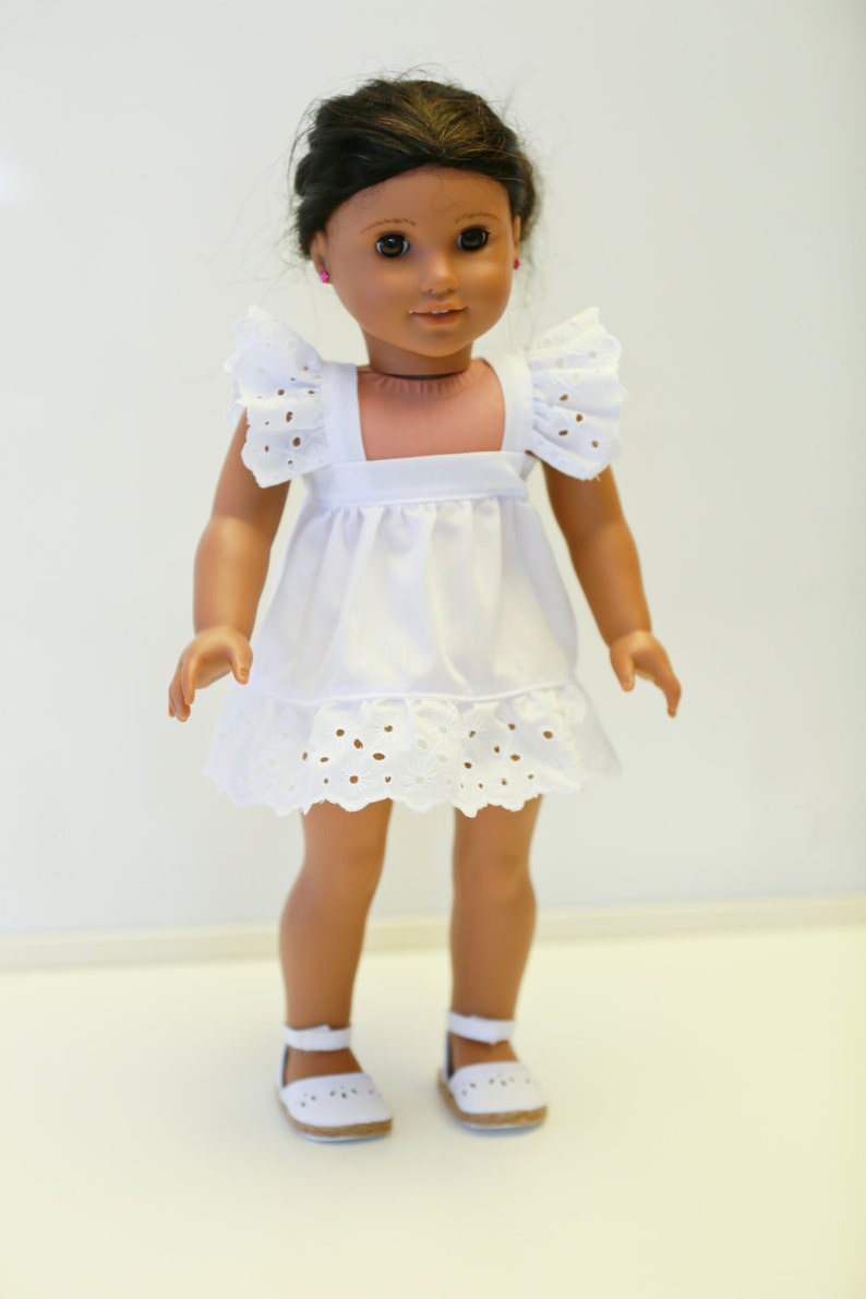 May include: A white doll with brown hair and brown eyes wearing a white eyelet dress and white shoes.