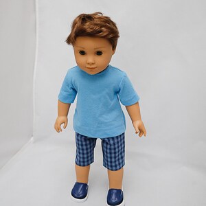 May include: A doll wearing a light blue t-shirt, blue and white checkered shorts, and dark blue shoes.