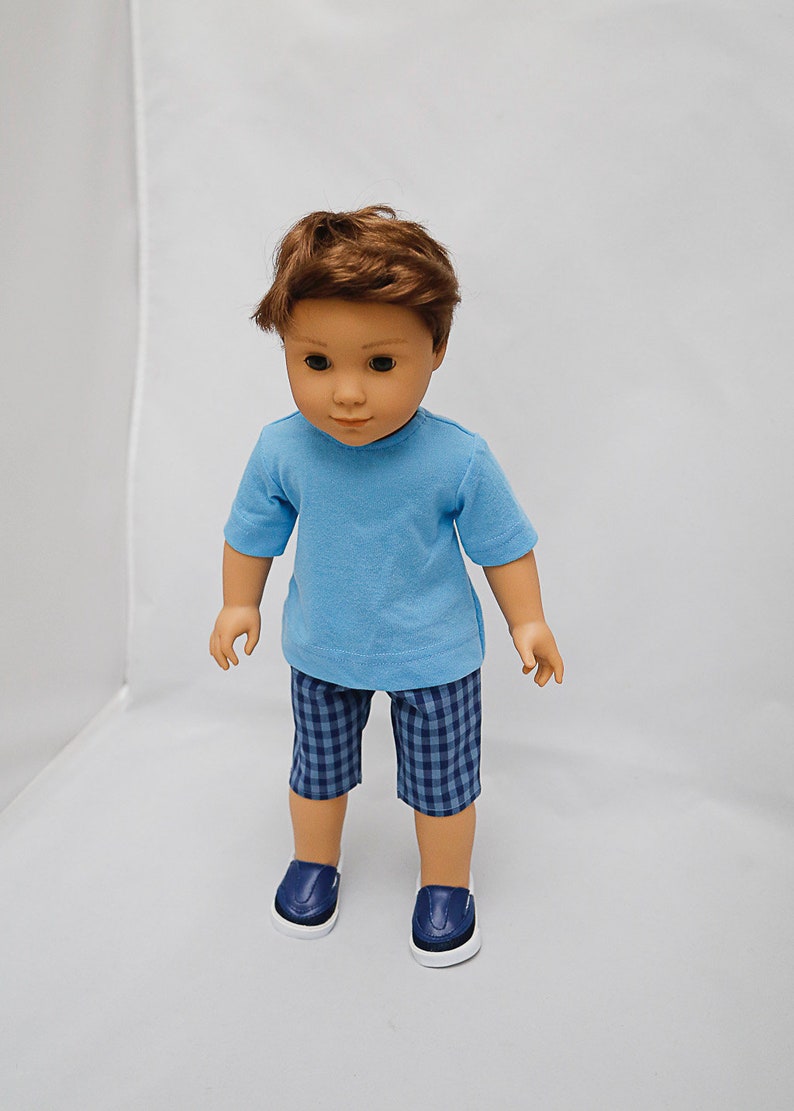 May include: A doll wearing a light blue t-shirt, blue and white checkered shorts, and dark blue shoes.
