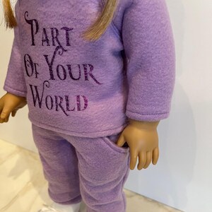 K&ouml;nnte beinhalten: Ein lilafarbenes Fleece-Puppenoutfit mit einem lang&auml;rmeligen Sweatshirt und einer passenden Hose. Das Sweatshirt hat den Text "Part of Your World" in lilafarbenem Glitzer.