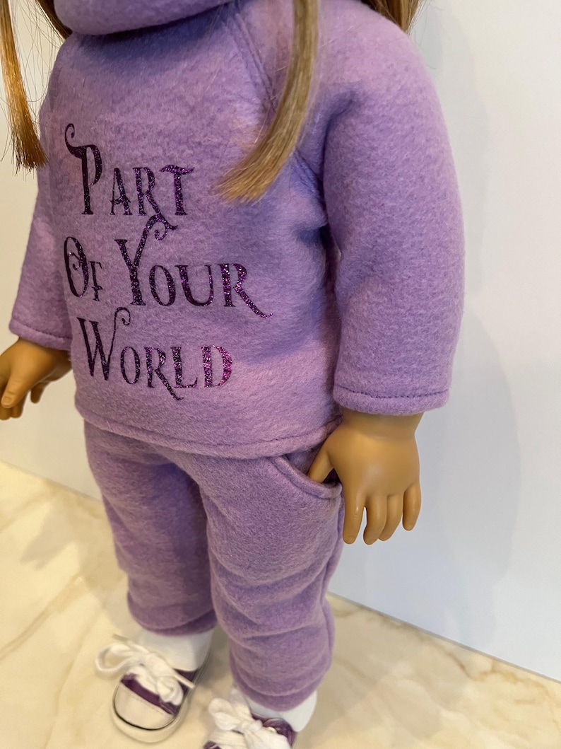 K&ouml;nnte beinhalten: Ein lilafarbenes Fleece-Puppenoutfit mit einem lang&auml;rmeligen Sweatshirt und einer passenden Hose. Das Sweatshirt hat den Text "Part of Your World" in lilafarbenem Glitzer.