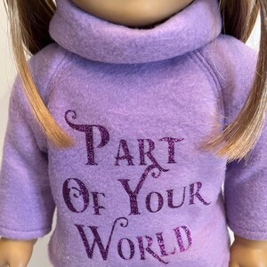 K&ouml;nnte beinhalten: Lilafarbener Fleece-Pyjama f&uuml;r eine Puppe mit dem Text "Part of Your World" in lilafarbenem Glitzer.